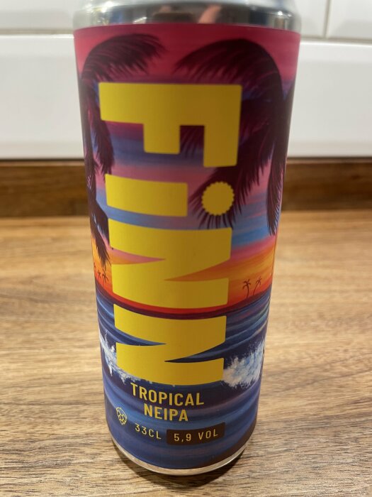 Burk med etiketten "FINN Tropical NEIPA, 33 CL, 5.9 VOL" mot en bakgrund med palmer, havsvågor och en solnedgång.