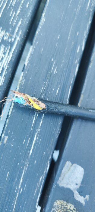 En skadad kabel med blottade ledningar, som ligger på ett mörkt träunderlag. Kabeln verkar ha fått sitt yttre hölje avskalat.