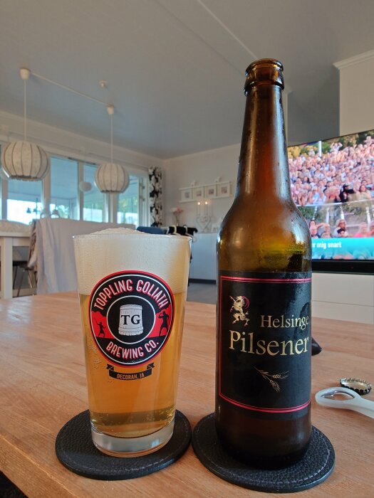 Ett glas öl med logotyp från Toppling Goliath Brewing Co. och en flaska Helsinge Pilsener står på ett bord i ett vardagsrum med en TV i bakgrunden.