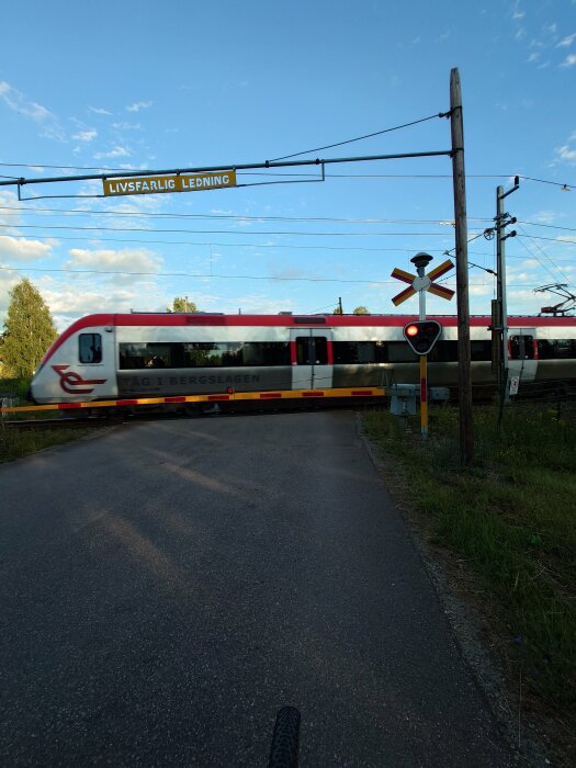 Tåg passerar en obevakad järnvägsövergång med ett varningsljus, åtföljt av en skylt som varnar för livsfarlig ledning ovan.