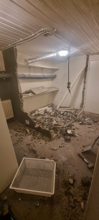 En källare under renovering med nedslagen vägg, avlägsnat skräp och exponerade rör längs väggar och tak. Verktyg och en lampa är synliga.