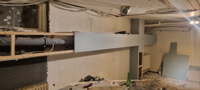 Bilden visar en källarvägg under renovering med synliga rör och halvinstallerade gipsskivor. Verktyg och byggmaterial ligger utspridda på golvet.