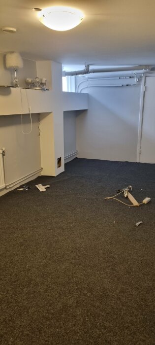 Nyinstallerad matta i ett 7 gånger 4 meter stort rum, under renovering med verktyg och material på golvet samt oskyddade rör och kablar.