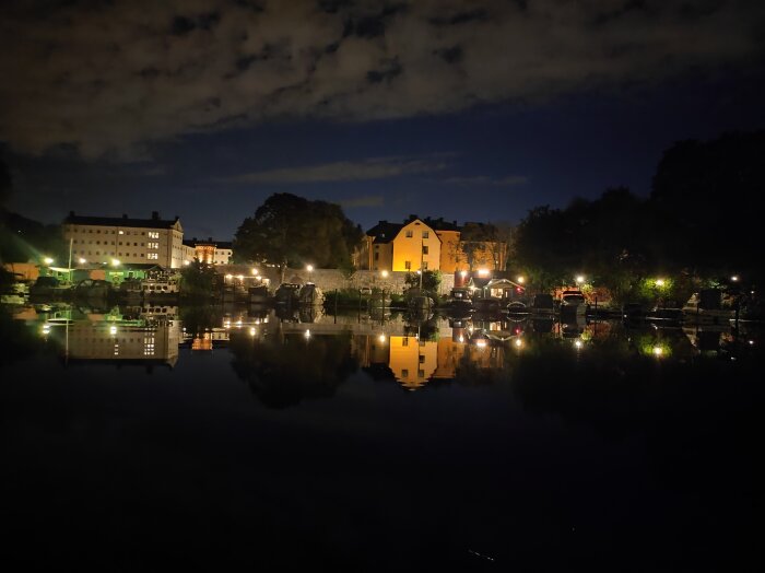 Nattvy över Långholmskanalen med upplysta byggnader och båtar som speglar sig i det stilla vattnet under en klar natthimmel.