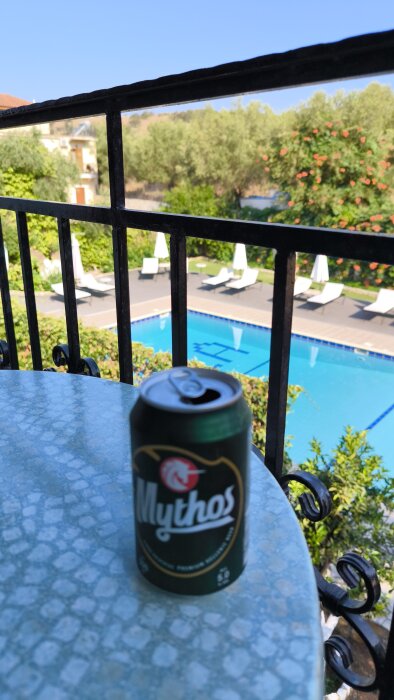 En öppnad burk Mythos-öl på ett bord med en pool, solstolar och grönska i bakgrunden.