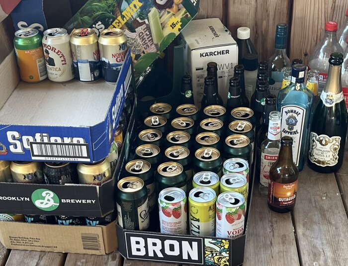 Flera kartonger och flaskor med öl, sprit och läsk på en träyta, inklusive märken som Sofiero och Bombay Sapphire, samt kartonger från Brooklyn Brewery.