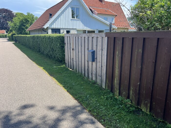 Trästaket i två färger längs trottoar bredvid vit husfasad och grön häck i villaområde, solig dag.