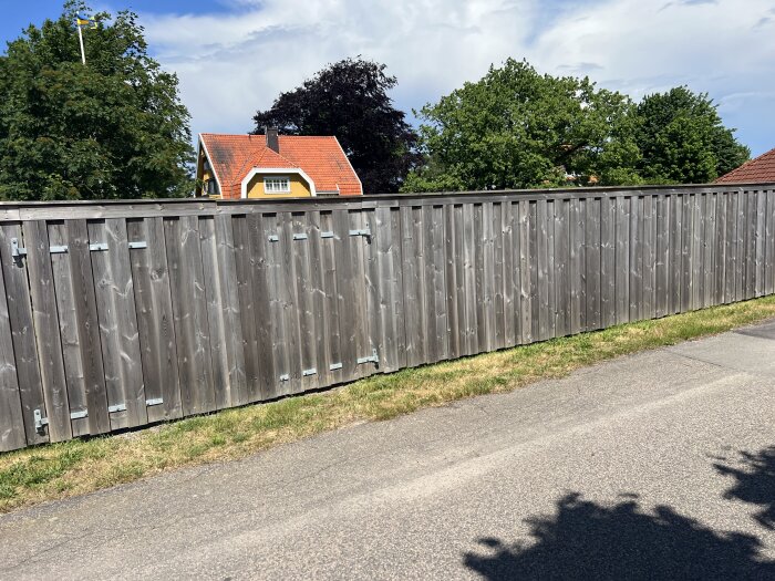 Högt träplank utomhus längs en asfalterad väg med gräsyta framför och hus och träd i bakgrunden under en delvis molnig himmel.