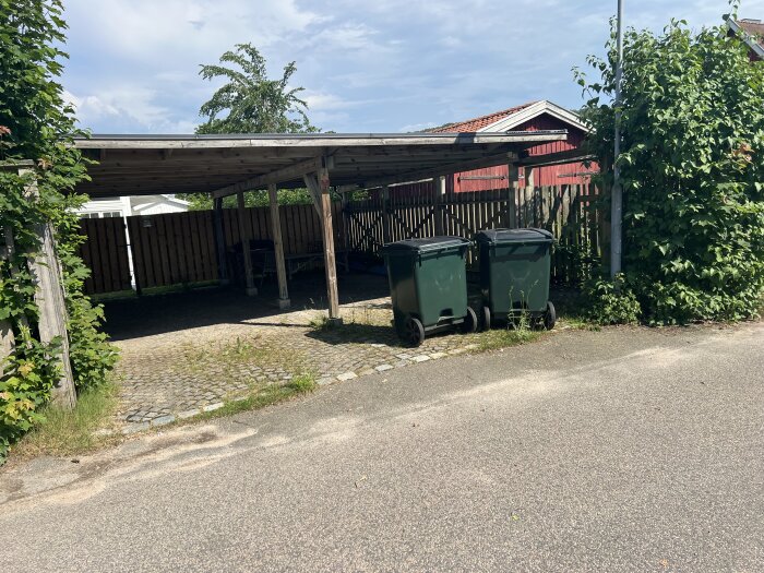 Carport med två gröna soptunnor och staket i bakgrunden. Staketet verkar vara av trä och är högre än 1,2 meter och ogenomsiktligt.