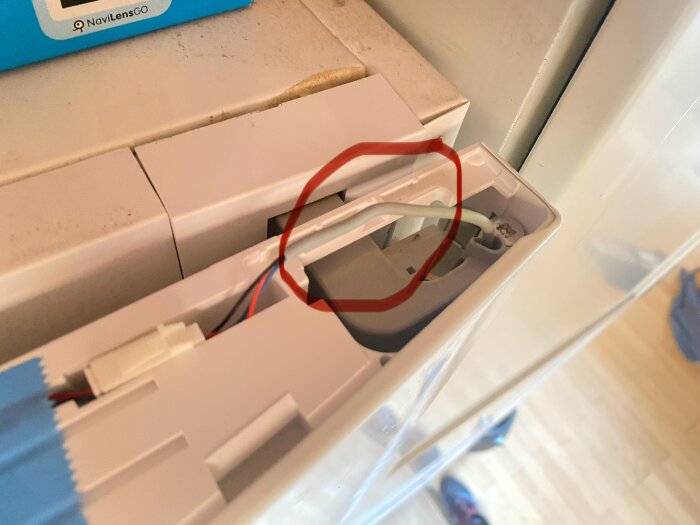 Öppen kylskåpsdörrs gångjärn med en lös kabel, omgiven av ett plastlock som delvis har tagits bort för att visa problemet.