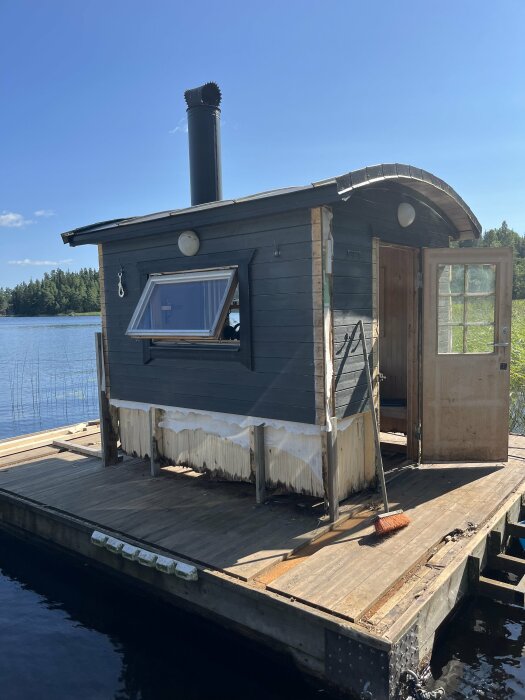 Bastuflotte vid sjö med rötskadad trall och nedre väggar. Dörr öppen, sopkvast på golvet, fällt fönster, synliga vattenskador runt om. Skog och vatten i bakgrunden.