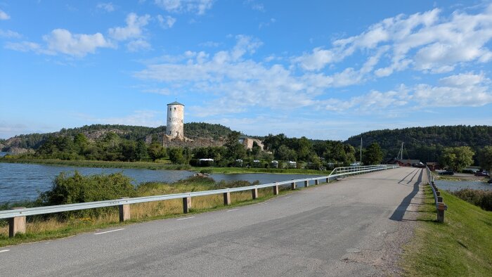 Bild av en vägbron över en vattendrag med en stenbyggd borg och ett runt torn på kullen i bakgrunden, omgivet av gröna träd och blommor under en klar blå himmel.