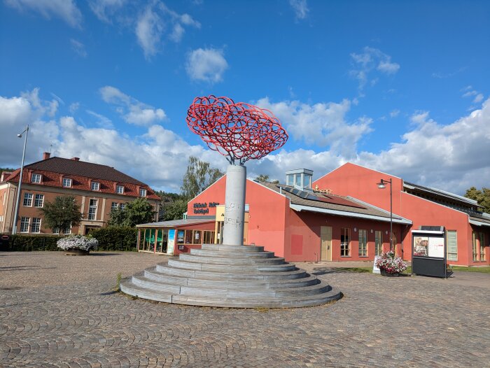 Ett modernt konstverk av rödlackerade metalltrådar ovanpå en betongpelare på ett torg framför en röd byggnad med skylten "Bibliotek, Museum, Kulturhuys".