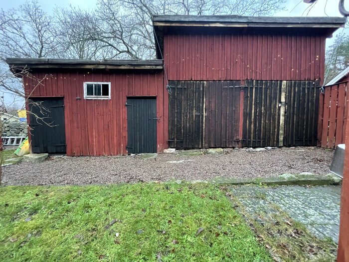 Rött uthus med två svarta dörrar och en trädgård framför, beläget i skogskanten med träd runt omkring.