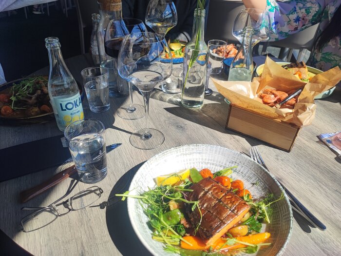 Lunchbord med dubbelrökt röding, rökta räkor, grillad biff och lax. Flaskor med mineralvatten, glas med drycker och en penna på bordet.