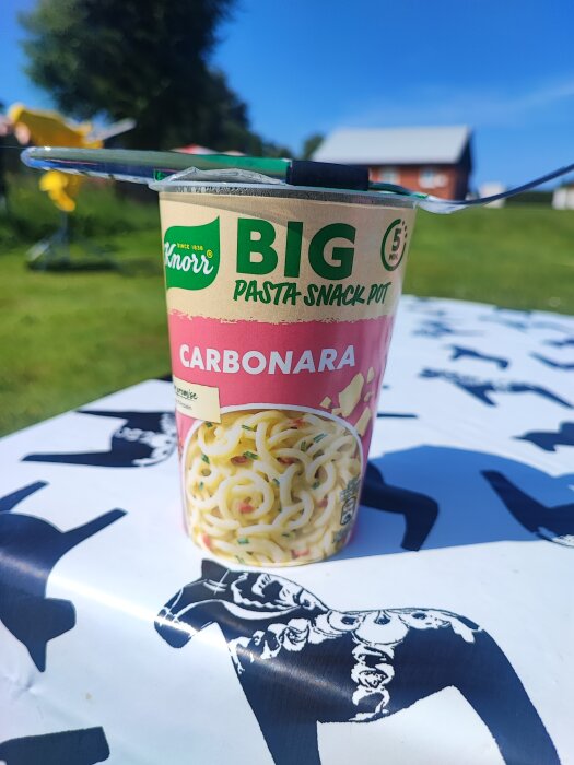 En kopp Knorr Big Pasta Snack Pot Carbonara på ett bord utomhus vid en campingplats, med en grönskande bakgrund och en blå himmel.