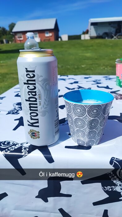 En ölburk Krombacher och en kaffemugg på ett bord med vit duk. I bakgrunden syns gräs, ett rött hus och en husvagn.