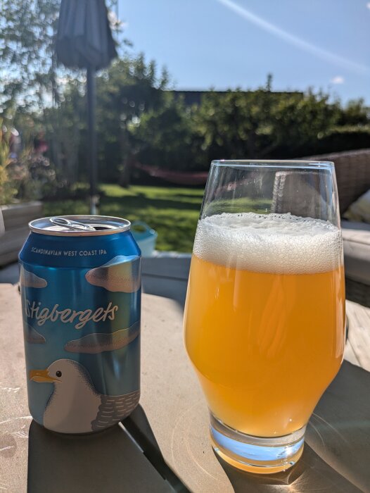 En burk Stigbergets Scandinavian West Coast IPA och ett fyllt glas med öl på ett bord utomhus, med en grönskande trädgård i bakgrunden.