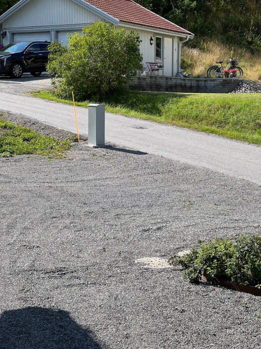 En modern brevlåda på en ställning står nära en grusad uppfart framför ett vitmålat hus med rött tak, svart bil parkerad och cyklar på terrassen.