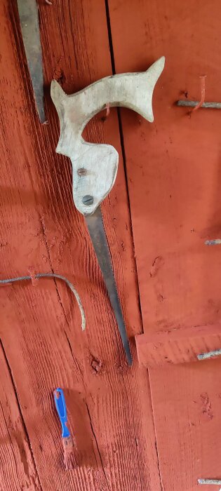 En liten handsåg med trähandtag hänger på en röd trävägg tillsammans med en krok och en blå pensel.