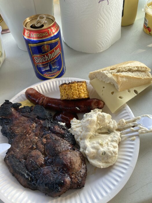 En papptallrik med grillat kött, korv, majskolv, potatissallad, bröd och ost. I bakgrunden syns en burk Stockholm Festival Beer.