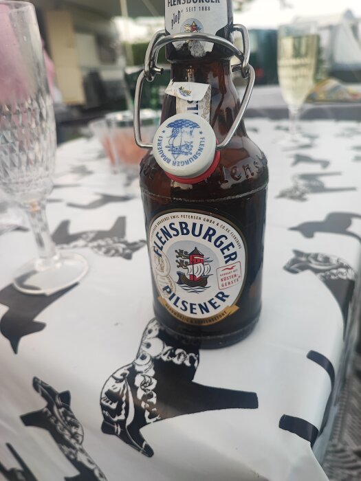 Flaska med Flensburger Pilsener på ett bord med vit duk med svarta mönster.