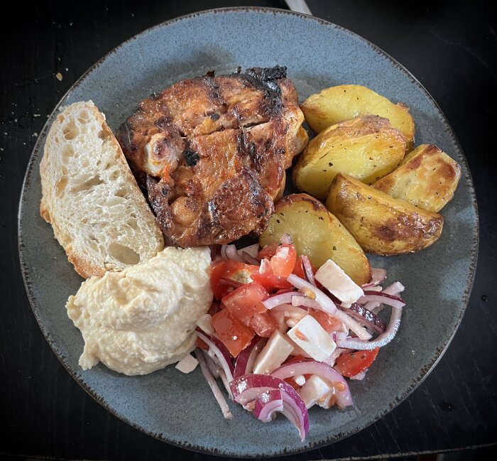 En tallrik med grillade kycklingben, hummus, tomat- och fetaostsallad, ugnstekt potatis, en bit bröd och en klick bearnaisesås.