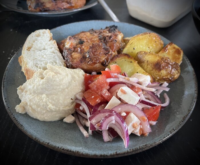 Tallrik med grillade kycklingben, hummus, tomat- och fetaostsallad, ugnsstekt potatis och en skiva bröd.