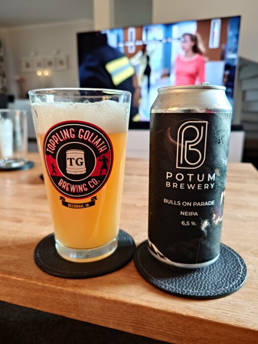 Glas med öl från Toppling Goliath Brewing Co. och en burk Potum Brewery Bulls on Parade NEIPA, 6,5 %, placerade på ett bord framför en TV.