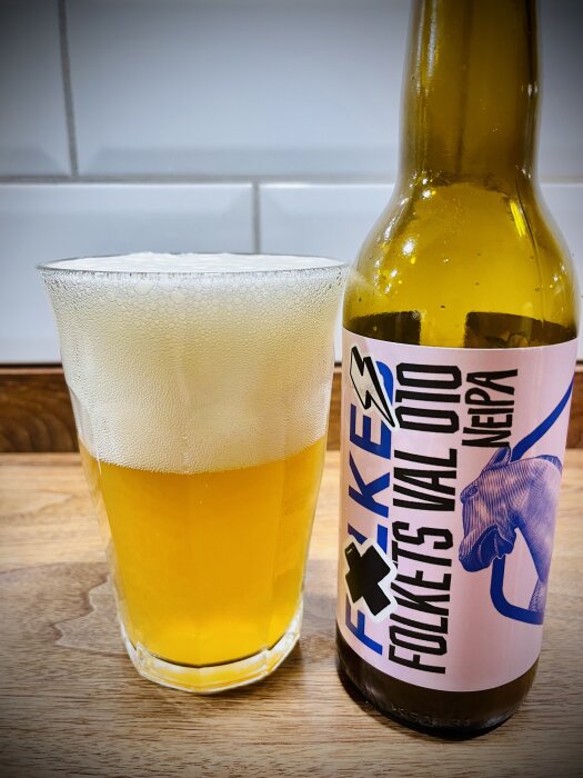 En flaska öl med etiketten "Folkets Val Otto NEIPA" och ett glas med skummande öl står på ett träbord.