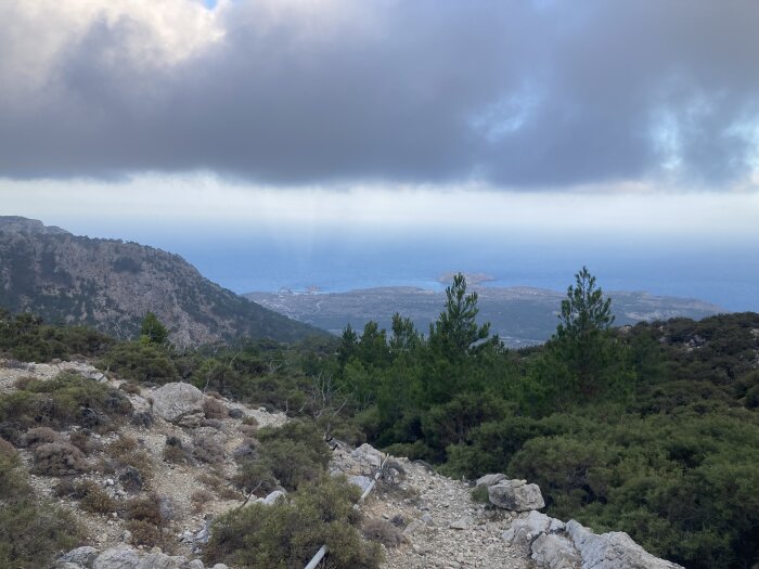 Utsikt från ett berg över ett hav med en molnig himmel, grönska och träd i förgrunden samt klippor och stenig mark.