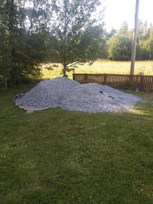 En stor hög med grus ligger på en presenning utomhus, omgiven av gräs, träd och ett trästaket i bakgrunden.