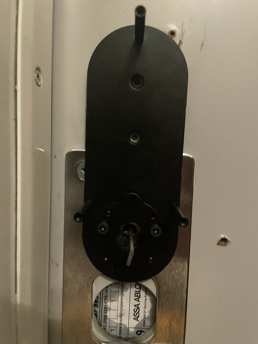 Närbild av en dörrlåsinstallation där en metallplatta och svart vredmedbringare monterats på insidan av en dörr med etikett "ASSA ABLOY" synlig.