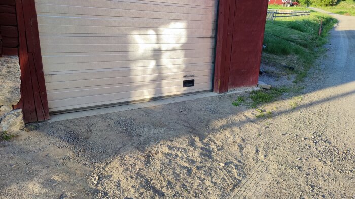 Garagedörren på en byggnad med sprucken träpanel på sidan och en gjuten betongtröskel. Grusig mark framför dörren och en grusväg bredvid.