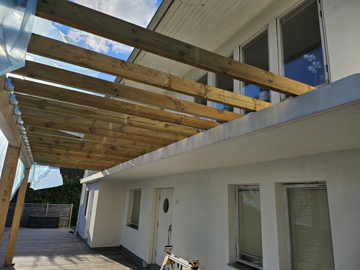 Byggnad med en pergola av träbjälkar på baksidan, fäst med balkskor. Blå presenning är delvis täckt över pergolan, och ett trästaket syns i bakgrunden.