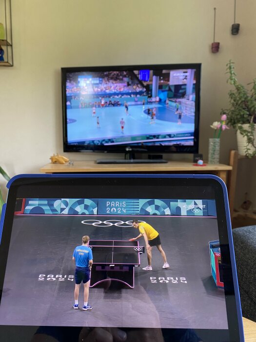 TV och surfplatta visar två olika sportevenemang från Paris 2024. På TV visas en handbollsmatch och på surfplattan en bordtennismatch.