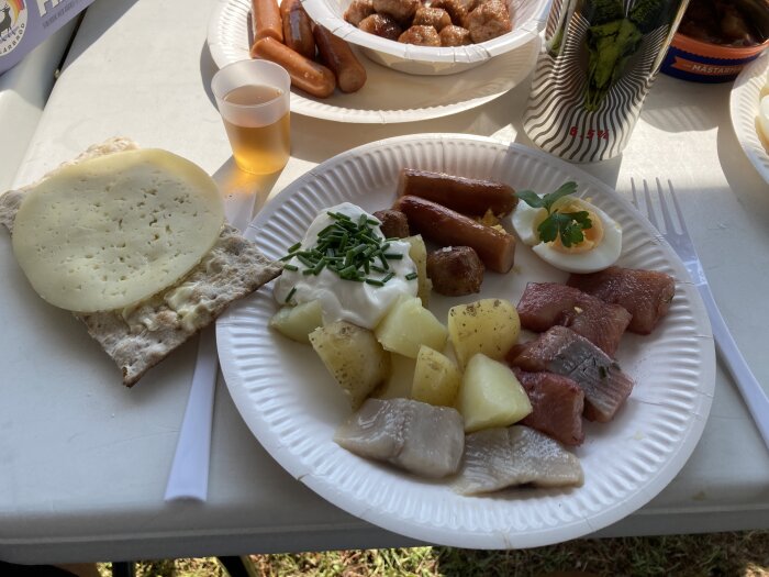 Pappersassiett med sillunch på ett bord: potatis, sillbitar, korv, ägg med dill, gräddfil med gräslök, knäckebröd med ost, dryck i plastglas och burk i bakgrunden.