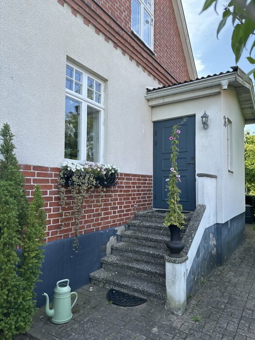 En äldre entré med en sliten trappa av betong framför ett hus från 1924 med tegelväggar och putsad fasad. Blomkrukor och grön växt syns vid trappan.