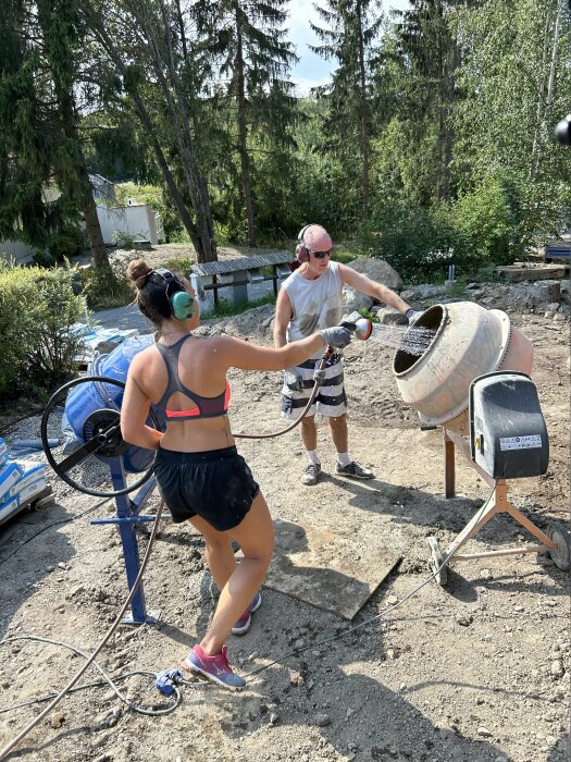 En man och en kvinna använder en cementblandare utomhus på en byggarbetsplats, omgivna av träd och byggmaterial.