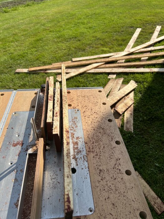 Brädor behandlas med en Bosch POF 1400 ACE fräs maskin på ett fräsbord utomhus, omgivna av gräsmatta.