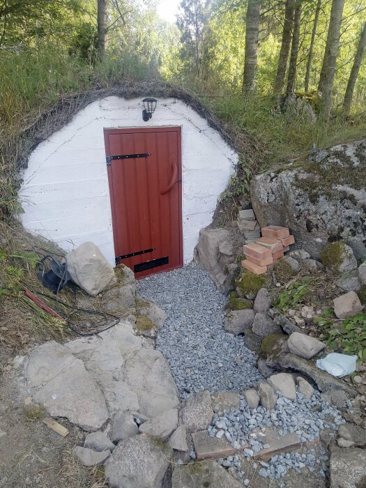 En röd dörr med ventil i nederkant och solcellsdriven ytterbelysning ovanpå, omgiven av vit puts och natursten. En gruslagd gång leder till dörren.