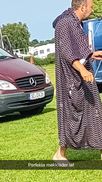 En man iklädd svartvitt randiga kläder står utomhus bredvid en röd Mercedes skåpbil, med husvagnar i bakgrunden.