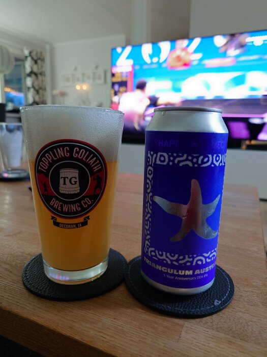 Ett glas öl från Toppling Goliath Brewing Co. och en ölburk med motiv av en sjöstjärna från Shapes & Objects Triangulum Australe 1 Year Anniversary DDH IPA framför en påslagen TV.