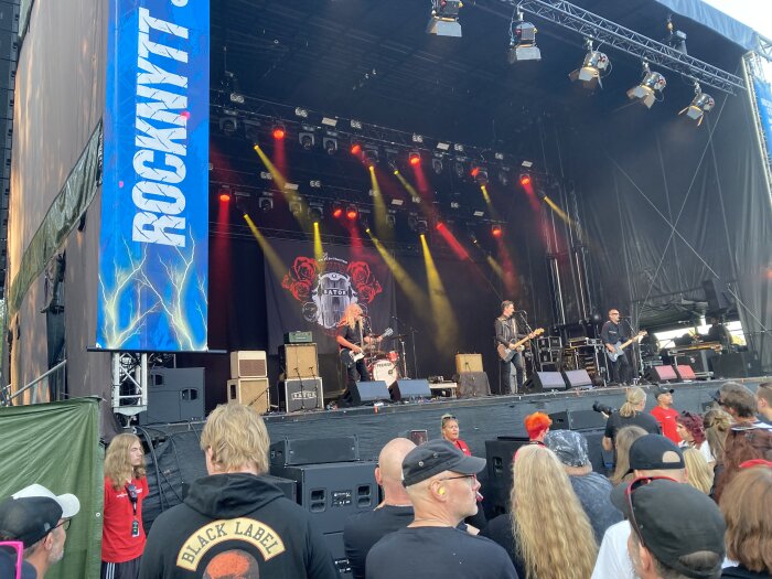 Rockband uppträder på en scen under "Rocknytt" festival, med en publik som tittar på. Ljuseffekter och bandets backdrop med rosor syns i bakgrunden.