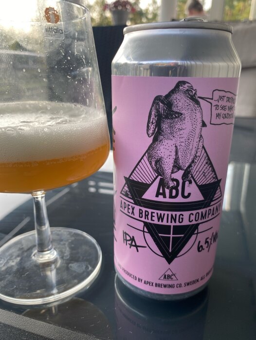 En burk Apex Brewing Company IPA med rosa etikett och skumfylld ölglas på ett bord i bakgrund.