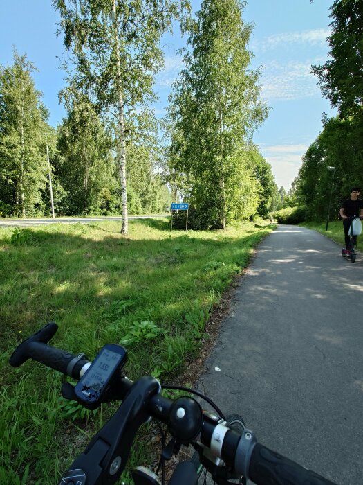 Cykelstyre på en asfalterad väg genom skog med skylten "Krylbo", person på elscooter i bakgrunden.