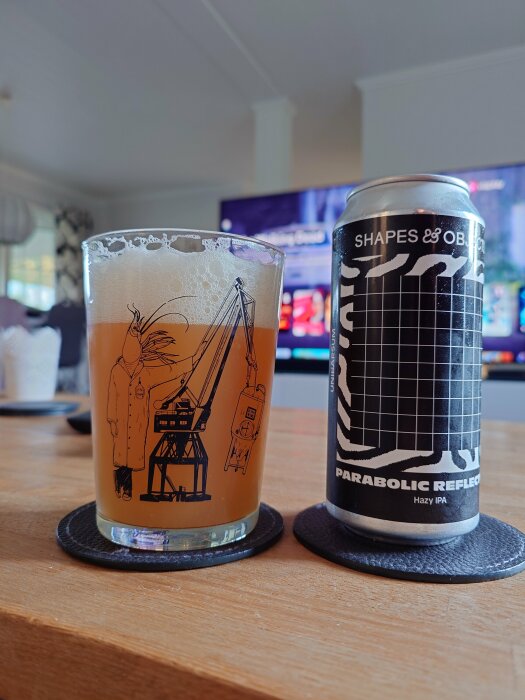 Ett glas med öl och en burk Shapes & Objects Parabolic Reflector Hazy IPA, stående på ett bord i ett vardagsrum med en TV i bakgrunden.