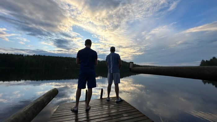 Två personer står på en brygga med ryggen mot kameran och ser ut över en stilla sjö under en molnig himmel vid solnedgång.