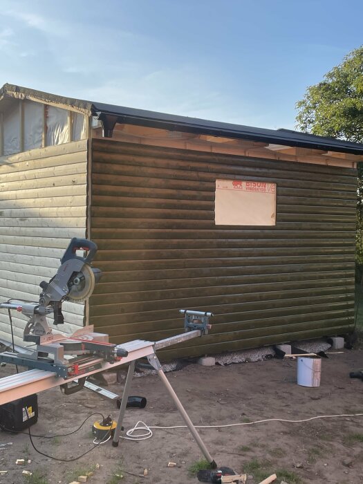 En nybyggd vägg med liggande träpanel som har blivit tjärbehandlad. En kap- och gersåg står på ett arbetsbord framför väggen.