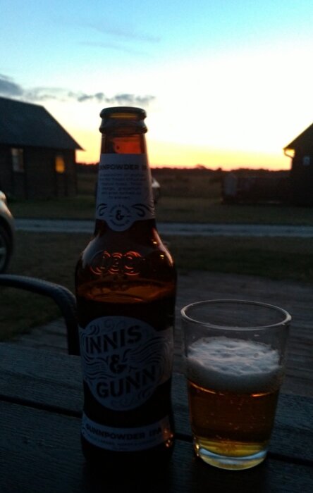 En flaska Innis & Gunn Gunnpowder IPA och ett glas öl på ett bord utomhus vid solnedgången, med stugor i bakgrunden.